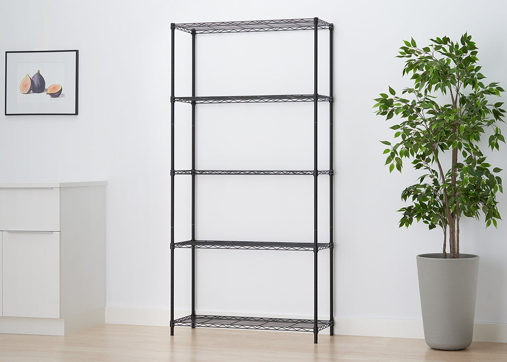 A black wire shelving unit stands empty in a brightly lit room next to a potted Ficus tree and a white sideboard, featuring five adjustable wire shelves. A framed art print of figs hangs on the white wall.