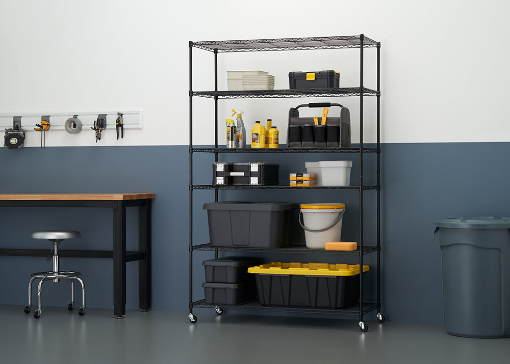 A tall, black wire shelving unit on wheels is fully stocked with various black and yellow storage containers, tools, and cleaning supplies in a clean, organized garage or workshop setting. The shelving unit is positioned against a two-toned wall (light gray above, dark blue below) next to a dark wood workbench with a metal stool.