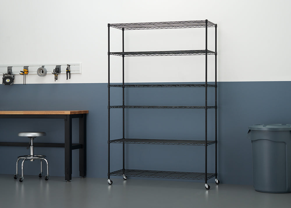 A black six-shelf wire shelving unit with wheels sits in a garage or workshop space. The background features a white and dark blue two-toned wall, a wooden workbench, a rolling metal stool, a tool rack, and a large gray trash can.