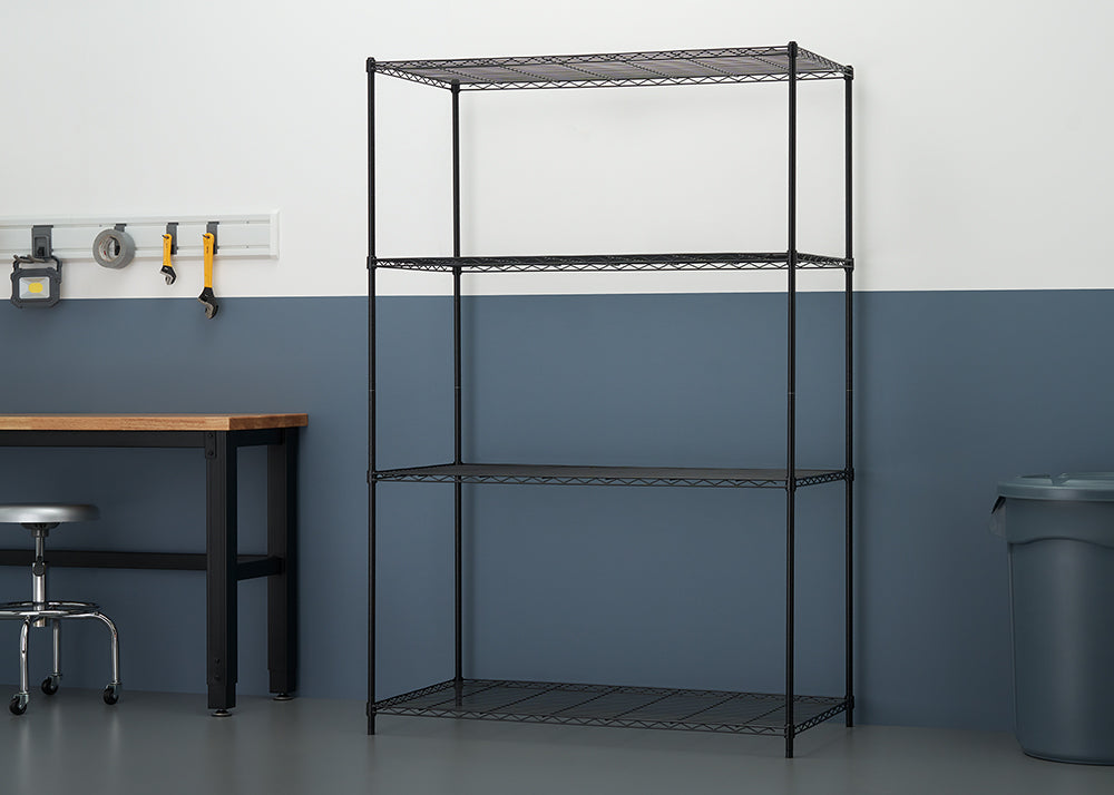 A four-tier black wire shelving unit is centered in a workshop or garage setting. The shelving is positioned against a wall painted white on top and a dark blue-grey on the bottom, with a workbench and a grey trash can visible on either side.