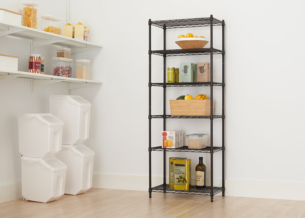 A tall, narrow, wire shelving unit is placed in a corner, stocked with various pantry items like produce, canned goods, boxed foods, and oils. To the left, there are white wall-mounted shelves and large plastic storage containers, suggesting this is a well-organized pantry or storage area.