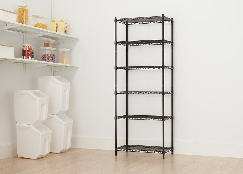 A tall wire shelving unit with five adjustable shelves stands against a white wall. The unit is in a room with light wood flooring, next to built-in white shelving on the left that holds clear and opaque storage containers.