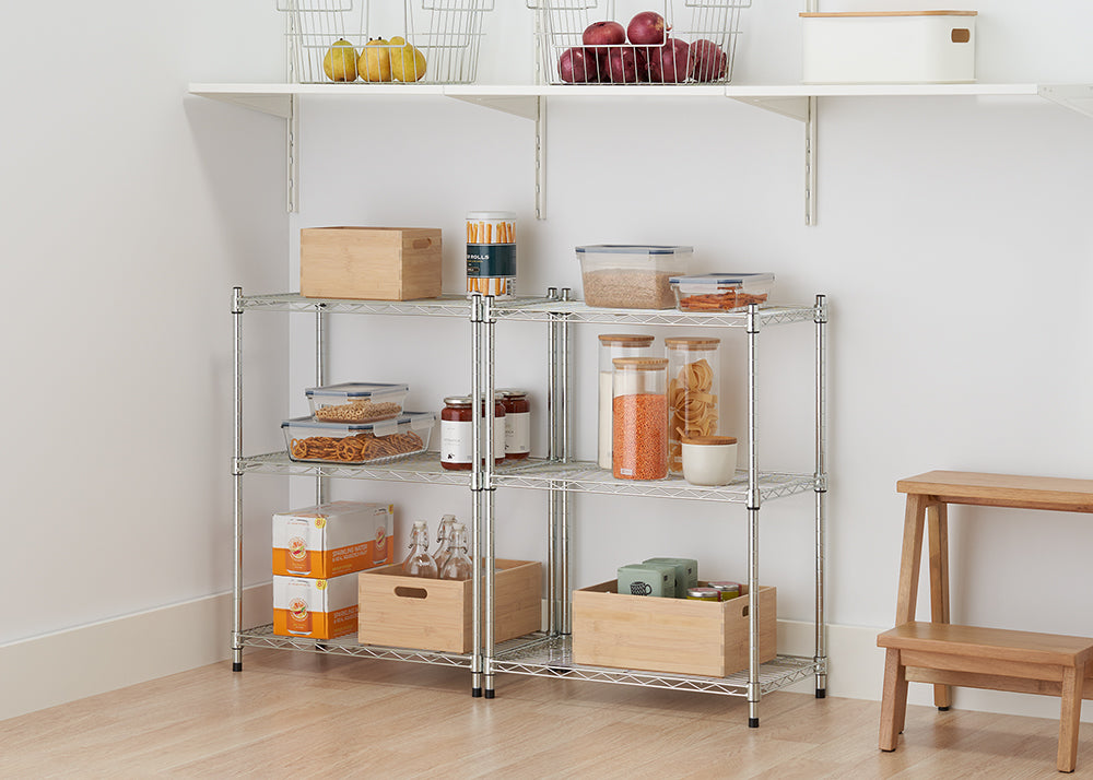 A low, three-tier chrome wire shelving unit holds various food items and storage containers in a pantry. Items on the shelves include glass jars of lentils and pasta, plastic containers of pretzels and crackers, and two wooden crates.