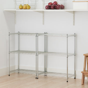 A two-unit chrome wire shelving system stands in the corner of a room, providing three tiers of storage. Above the units, a white wall-mounted shelf holds two wire baskets filled with fruit.