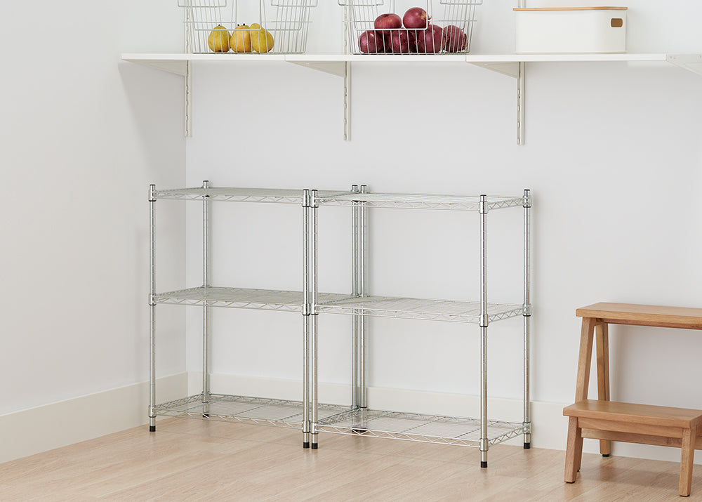 A two-unit chrome wire shelving system stands in the corner of a room, providing three tiers of storage. Above the units, a white wall-mounted shelf holds two wire baskets filled with fruit.