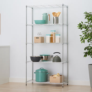 A five-shelf chrome wire rack stands in a kitchen setting, organized with various bowls, canisters, a tea kettle, and a large turquoise stockpot. The unit is positioned against a white wall between a white cabinet and a large potted plant, with a framed kitchen utensil print hanging to the left.