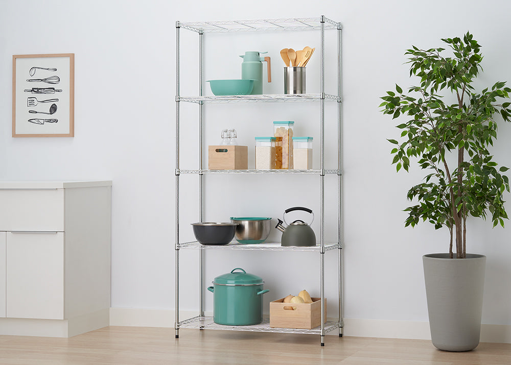 A five-shelf chrome wire rack stands in a kitchen setting, organized with various bowls, canisters, a tea kettle, and a large turquoise stockpot. The unit is positioned against a white wall between a white cabinet and a large potted plant, with a framed kitchen utensil print hanging to the left.