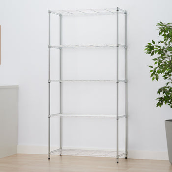 An empty five-shelf chrome wire rack is shown in a minimalist room setting against a white wall. The unit is flanked by a white cabinet and a framed print on the left, and a large potted plant on the right.