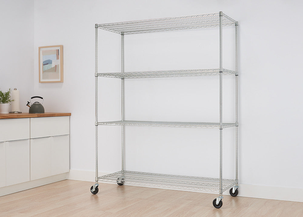 A silver metal wire shelving unit with four tiers stands against a white wall in a room. The unit has wheels at the bottom for mobility and is partially filled with items.