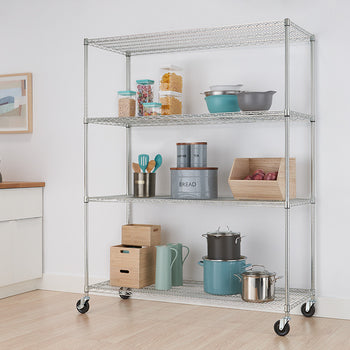 A silver metal wire shelving unit with four tiers and wheels stands in a kitchen, holding various pantry items and cookware. The kitchen features white cabinets, light wood countertops, and a light-colored floor.