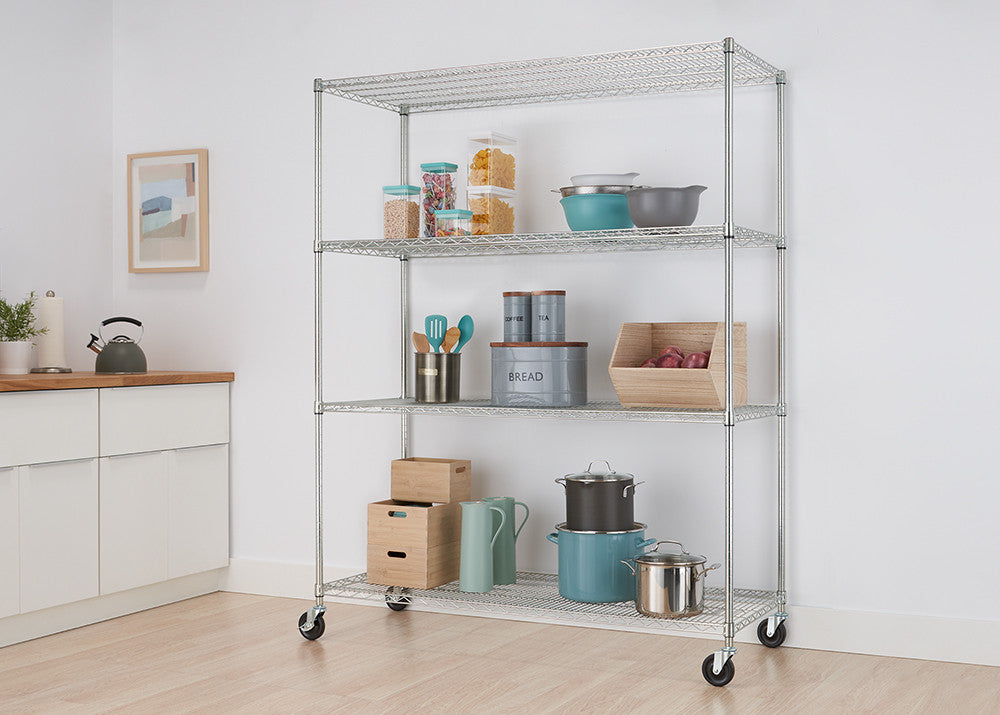 A silver metal wire shelving unit with four tiers and wheels stands in a kitchen, holding various pantry items and cookware. The kitchen features white cabinets, light wood countertops, and a light-colored floor.