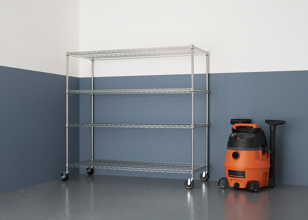 A chrome wire shelving unit with four tiers and wheels stands against a two-toned wall, next to an orange and black wet/dry vacuum. The shelving unit is empty and the floor is a glossy gray.
