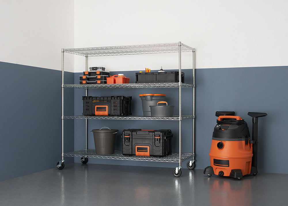 A 4-tier wire shelving unit with wheels, filled with various storage containers, stands against a two-tone wall with a glossy floor. An orange wet/dry vacuum is positioned to the right of the shelving unit.