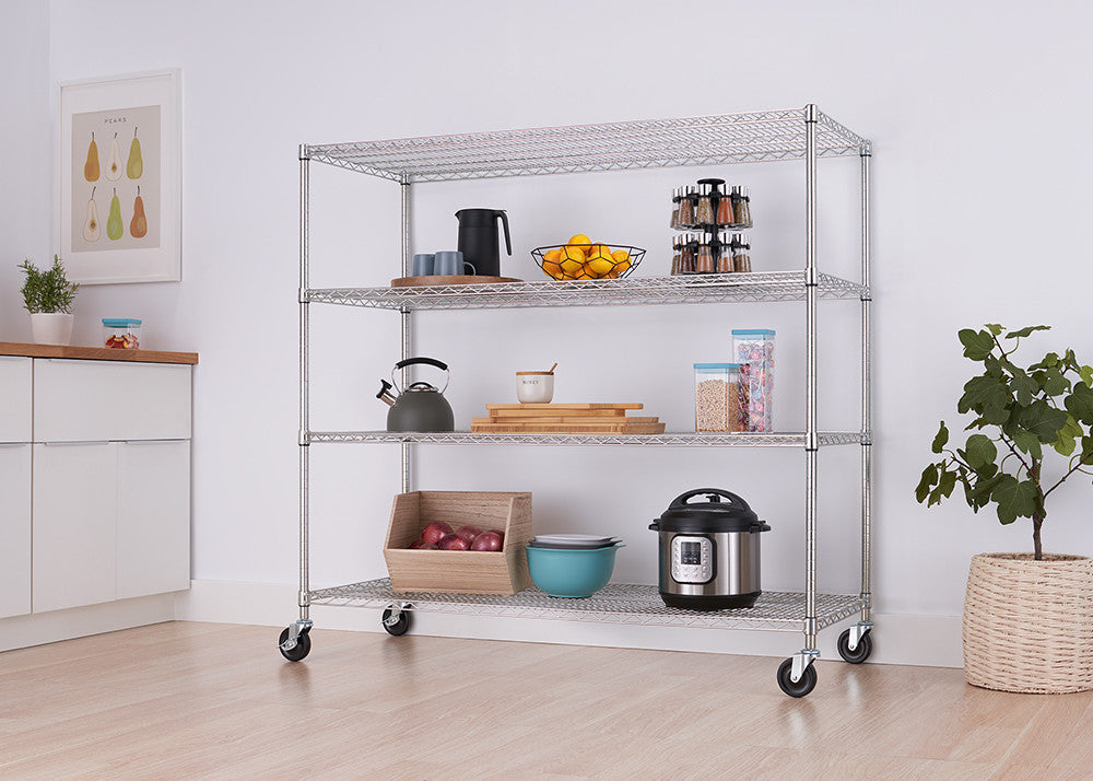 A four-tier wire shelving unit with wheels is shown in a bright, modern kitchen. The shelves hold various kitchen items, and the unit is positioned against a white wall with a framed picture and a potted plant nearby.