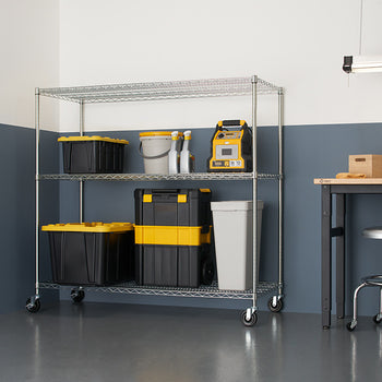 A silver wire shelving unit with three tiers is shown in a garage setting, holding various storage containers and tools. The shelving unit has wheels and sits on a gray floor against a two-tone gray and white wall.