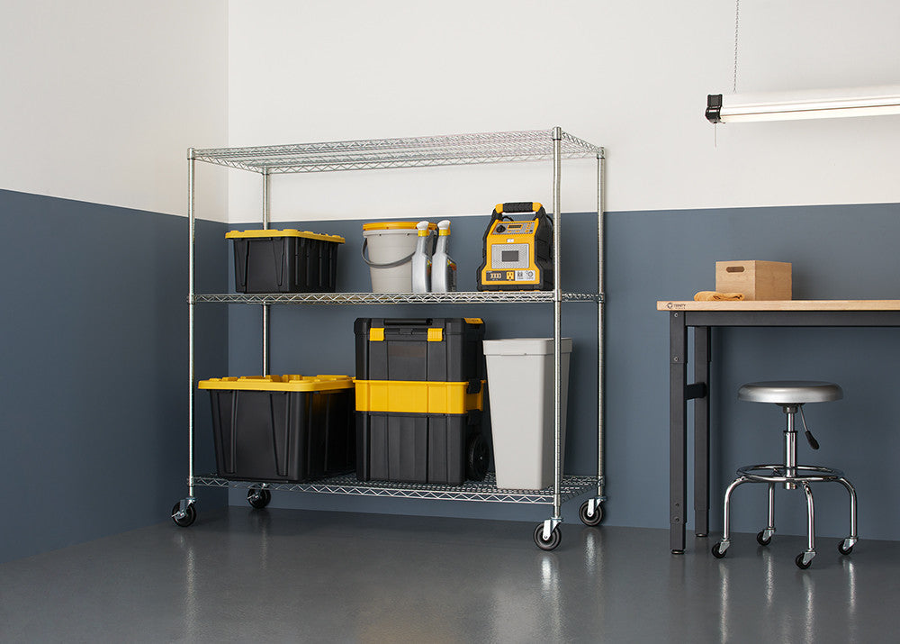A silver wire shelving unit with three tiers is shown in a garage setting, holding various storage containers and tools. The shelving unit has wheels and sits on a gray floor against a two-tone gray and white wall.