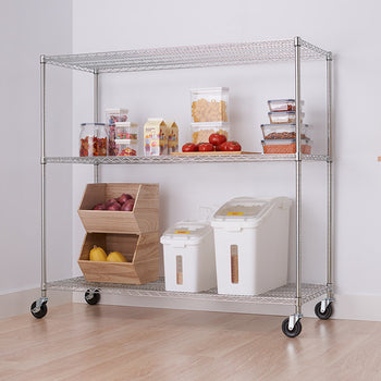 A silver metal wire shelving unit with four tiers and wheels holds various kitchen items like containers and produce. The unit is positioned against a white wall next to a white cabinet with a wooden top.