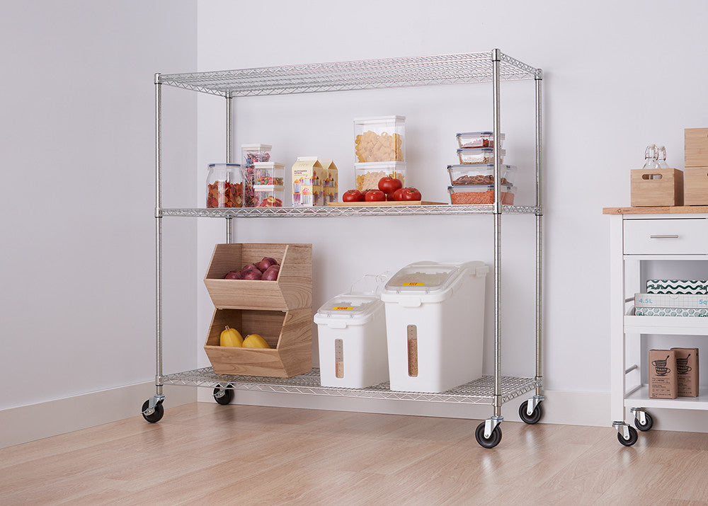 A silver metal wire shelving unit with four tiers and wheels holds various kitchen items like containers and produce. The unit is positioned against a white wall next to a white cabinet with a wooden top.