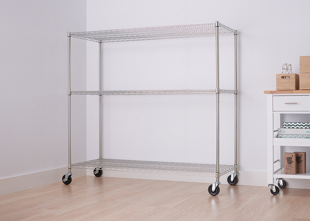 A three-tier wire shelving unit with wheels is shown in a room with light-colored walls and flooring. The unit is empty and positioned against a white wall, with a partial view of a white cart with a wooden top on the right.