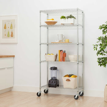 A silver chrome 5-tier wire shelving unit on caster wheels holds kitchen accessories, books, a blender, and storage bins. Set in a bright, modern room, this versatile rolling storage rack stands beside a potted plant and wooden flooring.