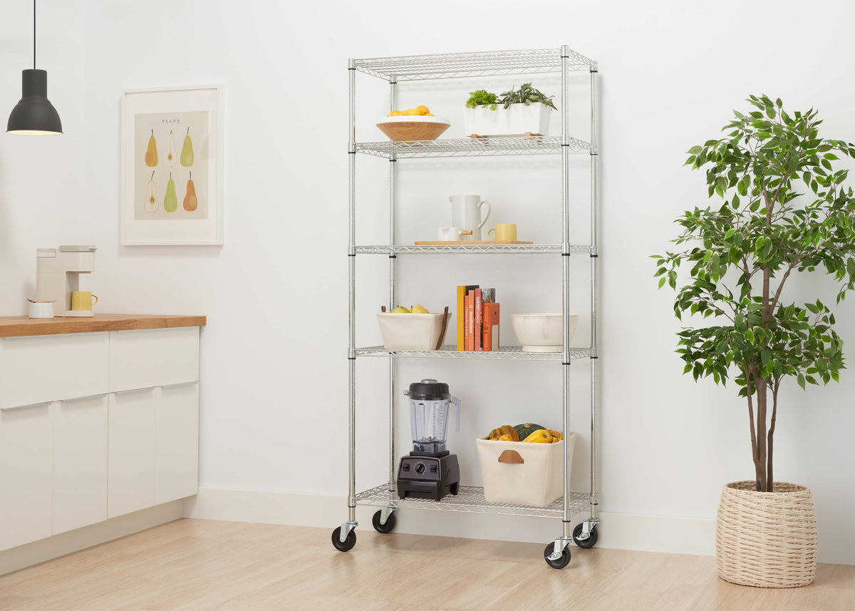 A silver chrome 5-tier wire shelving unit on caster wheels holds kitchen accessories, books, a blender, and storage bins. Set in a bright, modern room, this versatile rolling storage rack stands beside a potted plant and wooden flooring.