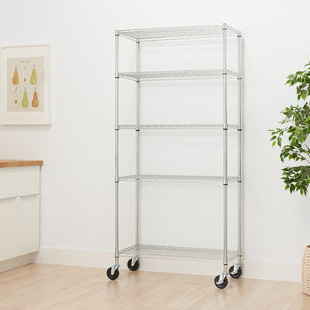 A five-tier chrome wire shelving unit stands against a white wall. The unit has wheels on the bottom and is shown in a room with a plant, a framed picture of pears, and a kitchen counter.