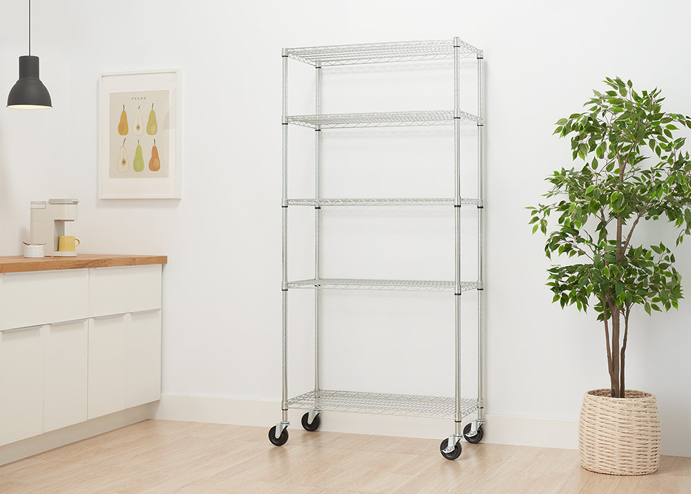 A five-tier chrome wire shelving unit stands against a white wall. The unit has wheels on the bottom and is shown in a room with a plant, a framed picture of pears, and a kitchen counter.