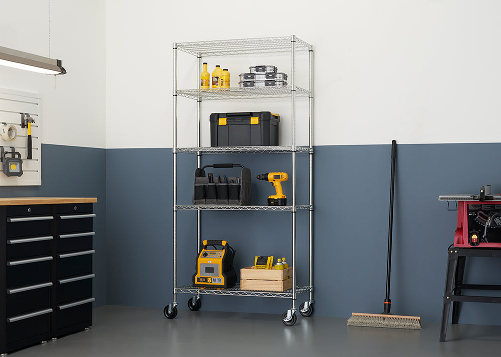 A chrome wire shelving unit with four shelves sits in a garage. The shelves are filled with tools and supplies, including a yellow and black toolbox, a tool bag, a cordless drill, a wooden crate with bottles, and a battery jumper box.