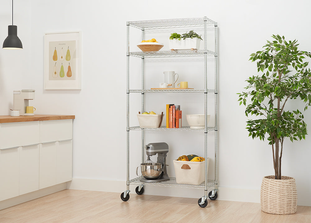 A five-tier wire shelving unit with wheels is filled with various items. The shelves hold a stand mixer, a large basket with gourds, books, bowls, and potted plants.