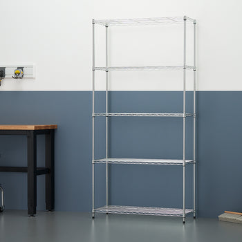 A five-tier chrome wire shelving unit stands empty in a workshop against a two-tone white and blue wall. To the left, a workbench with tools and a black rolling stool are visible, while a push broom and trash can sit to the right.