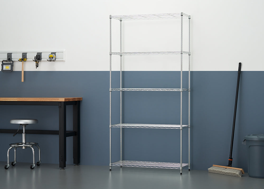 A five-tier chrome wire shelving unit stands empty in a workshop against a two-tone white and blue wall. To the left, a workbench with tools and a black rolling stool are visible, while a push broom and trash can sit to the right.