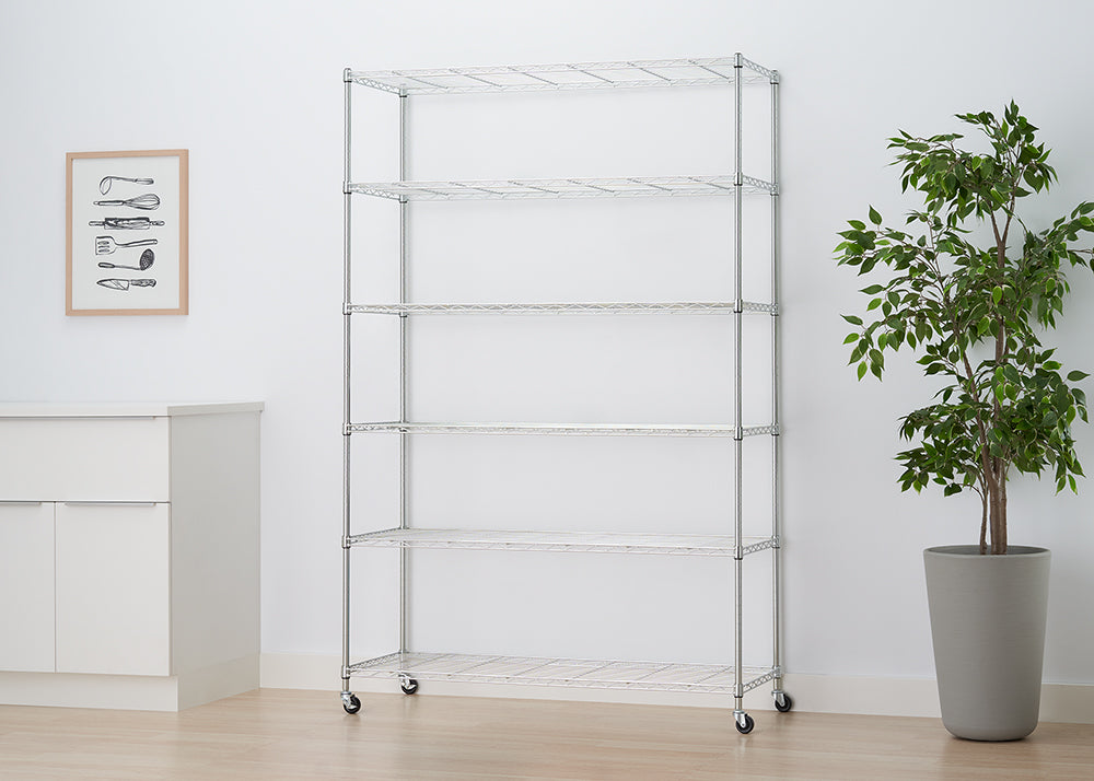 A six-shelf chrome wire rolling rack is shown centered against a white wall. To the right is a large potted plant, and to the left is a white cabinet with a framed print above it.