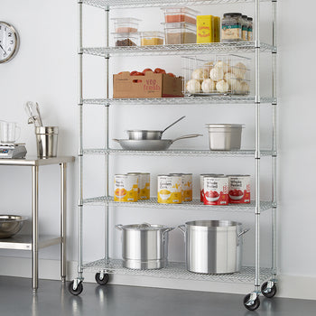 A six-tier chrome wire shelving unit on caster wheels, positioned against a plain white wall in a commercial kitchen setting. The shelves are organized with kitchen essentials and food supplies, including large stockpots, stacked canned goods, stainless steel pans, a crate of tomatoes, a wire basket of onions, and various clear food storage containers.
