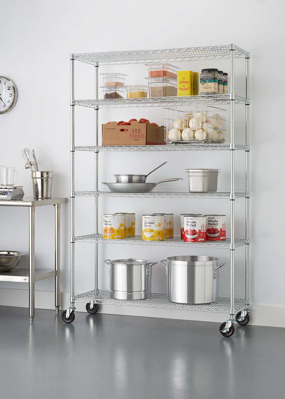 A six-tier chrome wire shelving unit on caster wheels, positioned against a plain white wall in a commercial kitchen setting. The shelves are organized with kitchen essentials and food supplies, including large stockpots, stacked canned goods, stainless steel pans, a crate of tomatoes, a wire basket of onions, and various clear food storage containers.
