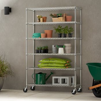 A six-tier chrome wire shelving unit on caster wheels, positioned against a textured grey wall in a patio or garage setting. The shelves are neatly organized with various gardening supplies, including potted plants, terracotta pots, bags of soil, and a watering can, while a green wheelbarrow sits nearby.