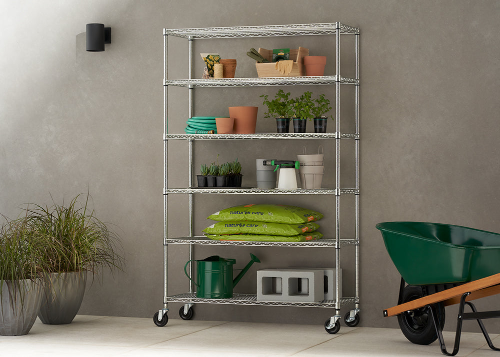 A six-tier chrome wire shelving unit on caster wheels, positioned against a textured grey wall in a patio or garage setting. The shelves are neatly organized with various gardening supplies, including potted plants, terracotta pots, bags of soil, and a watering can, while a green wheelbarrow sits nearby.
