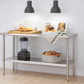 A stainless steel commercial work table features a large wooden cutting board with halved butternut squash, prep bowls, and a cookbook illuminated by two overhead black pendant lights. The bottom storage shelf conveniently holds a countertop blender and a basket filled with onions against a white subway tile backdrop.