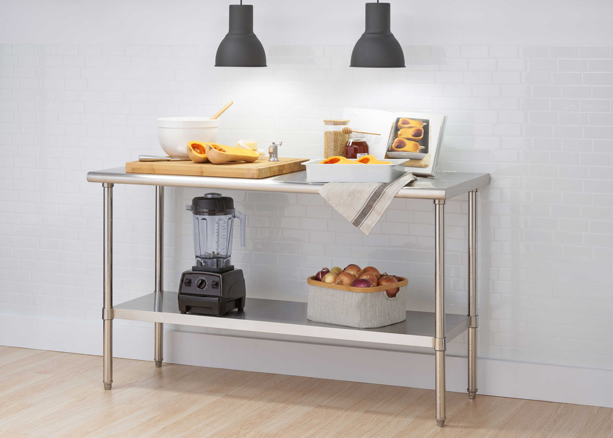 A stainless steel commercial work table features a large wooden cutting board with halved butternut squash, prep bowls, and a cookbook illuminated by two overhead black pendant lights. The bottom storage shelf conveniently holds a countertop blender and a basket filled with onions against a white subway tile backdrop.