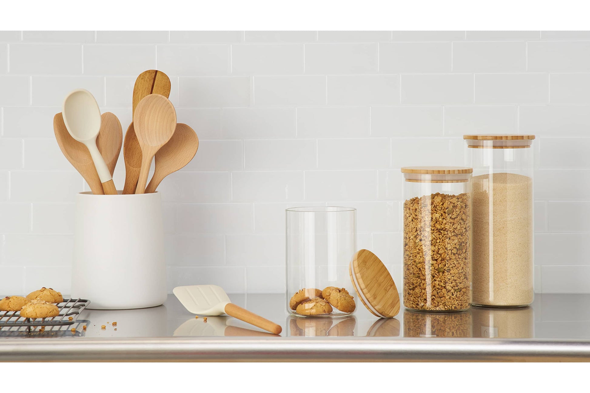 A kitchen countertop features a white utensil holder filled with wooden and white cooking spoons. To the right, three glass canisters with bamboo lids hold cookies, granola, and sugar, respectively, while more cookies sit on a cooling rack to the left.