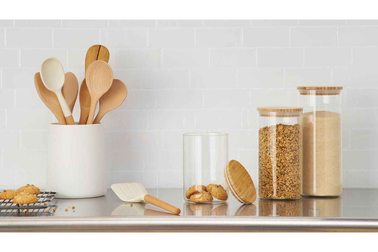 A kitchen countertop features a white utensil holder filled with wooden and white cooking spoons. To the right, three glass canisters with bamboo lids hold cookies, granola, and sugar, respectively, while more cookies sit on a cooling rack to the left.