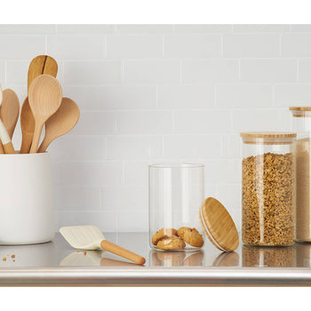 A kitchen countertop features a white utensil holder filled with wooden and white cooking spoons. To the right, three glass canisters with bamboo lids hold cookies, granola, and sugar, respectively, while more cookies sit on a cooling rack to the left.