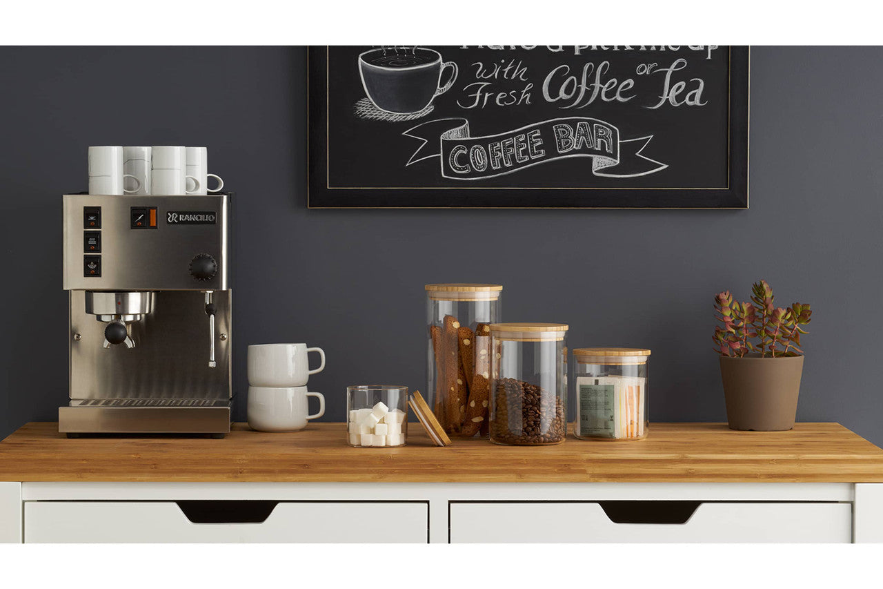 A coffee corner setup features a stainless steel espresso machine with stacked white mugs on top. Below it, there are stacked white mugs, a clear glass with sugar cubes, jars containing coffee beans and snacks, and a potted plant, all arranged on a light wooden countertop against a dark gray wall with a chalkboard sign that reads "Coffee Bar".