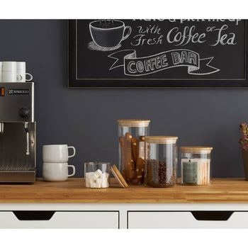 A coffee corner setup features a stainless steel espresso machine with stacked white mugs on top. Below it, there are stacked white mugs, a clear glass with sugar cubes, jars containing coffee beans and snacks, and a potted plant, all arranged on a light wooden countertop against a dark gray wall with a chalkboard sign that reads 