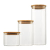 Three clear glass canisters of varying heights stand against a white background. Each canister has a round bamboo lid.