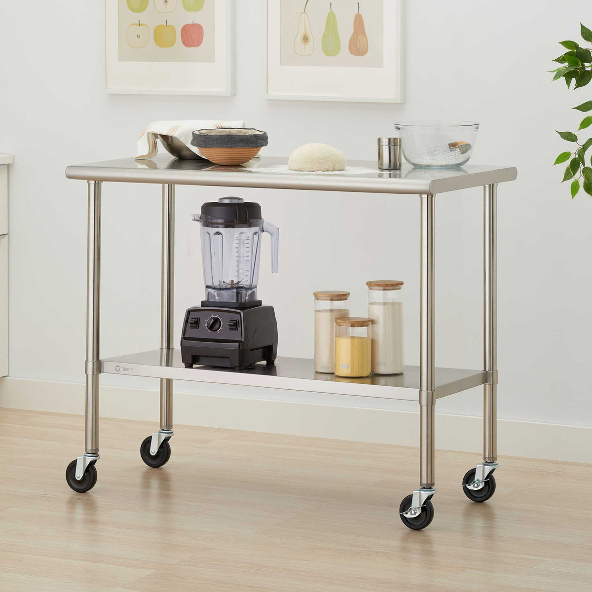 A two-tier stainless steel rolling kitchen cart holds a blender, glass storage jars, mixing bowls, and baking essentials. Its spacious shelves and four caster wheels provide convenient storage and mobility in a bright, modern kitchen.