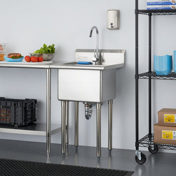 A stainless steel utility sink with a gooseneck faucet stands against a light gray wall in a commercial setting. To the left, a stainless steel table holds food items, and to the right, a black wire shelving unit displays various supplies.