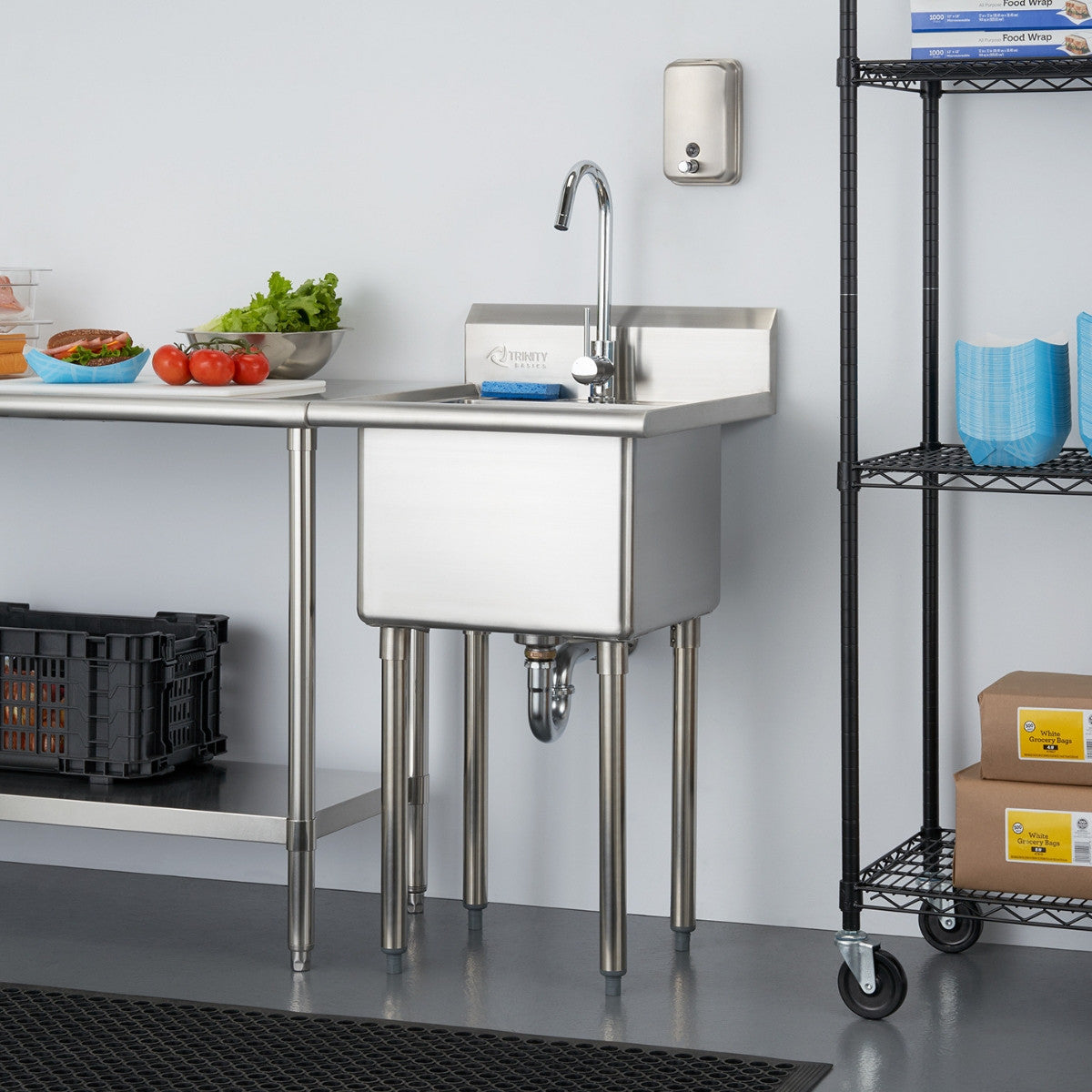 A stainless steel utility sink with a gooseneck faucet stands against a light gray wall in a commercial setting. To the left, a stainless steel table holds food items, and to the right, a black wire shelving unit displays various supplies.