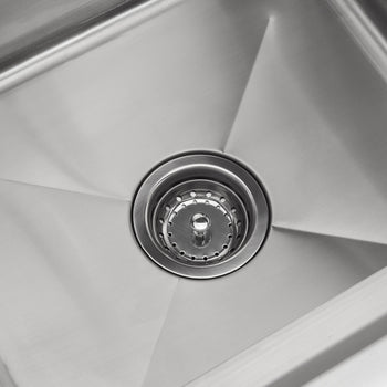 A close-up, overhead shot shows the bottom of a stainless steel kitchen sink. A round drain with small holes and a central post is visible in the center of the sink basin.