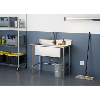 A stainless steel utility sink with a pull-out faucet stands against a blue and white tiled wall in a garage or utility room. To the left, a metal shelving unit holds various storage bins and cleaning supplies, while a broom leans against the wall to the right.