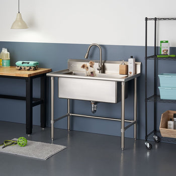 A small, fluffy, white and brown dog stands in a stainless steel utility sink with a gooseneck faucet. The sink is freestanding with tubular stainless steel legs and is positioned in a room with a two-toned wall, a wooden-topped table to the left, and a black wire shelving unit to the right.